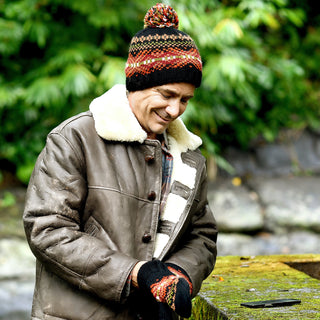 Wearing a brown shearling coat, matching gloves, and the Hand Knit Explore Hat—a wool beanie with a pom-pom—a person stands outdoors beside a mossy surface, framed by green foliage and stone steps in the background.