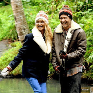 Two adults, dressed for cold weather in winter coats and gloves, stand smiling by a creek surrounded by green foliage and a tree trunk. Both are wearing cozy Hand Knit Explore Hats, enjoying the outdoors in their warm beanies.