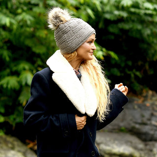 Wearing the Hand Knit Sena Hat, a person in a black coat with a large white collar stands outdoors in front of green foliage, looking to the side.