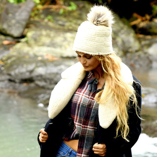 Wearing the Hand Knit Sena Hat, a person with long blonde hair stands outdoors near a rocky stream. They are also dressed in a plaid shirt and a coat with a large shearling collar, and the hat features a faux fur pompom.