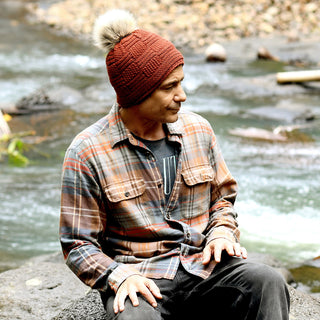 Wearing a plaid shirt and black pants, a person sits on a rock by a flowing creek, looking to the side and keeping warm in the cozy Hand Knit Sena Hat.