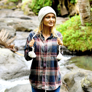 A woman stands by a rocky stream, holding the tassels of her Hand Knit Nest Hat. She is wearing a plaid shirt, surrounded by lush green plants and trees in the background.