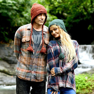 Two people in plaid shirts and knit hats, including the Hand Knit Nest Hat, stand close together outdoors by a waterfall and greenery. One person has their arm behind their back, while the other leans their head on their companion’s shoulder.