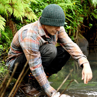 Wearing a plaid shirt, dark pants, boots, and the Hand Knit Grid hat, a person squats by the edge of a clear stream surrounded by green plants, touching the water with one hand and looking down.