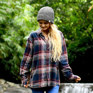 Wearing the Hand Knit Grid hat—a gray, handmade beanie—a person with long blonde hair, dressed in a plaid shirt and blue jeans, stands outdoors before a backdrop of green foliage, looking down as they hold the shirt’s sleeves.