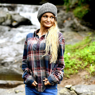 A woman with long blonde hair, wearing the Hand Knit Grid hat in gray Merino wool and a plaid flannel shirt, stands outdoors near a rocky stream surrounded by greenery, looking to the side with her hands in her pockets.