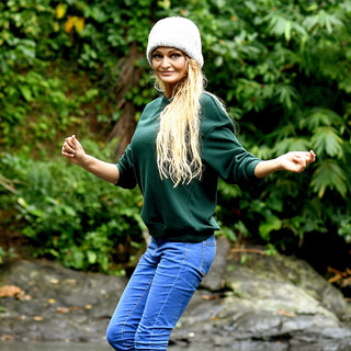 Wearing the Hand Knit Grid hat in gray, a woman stands outdoors on a rocky surface surrounded by lush green foliage. She smiles warmly, dressed in a green sweater and blue jeans, with her arms raised and elbows bent.