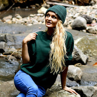 A woman with long blonde hair wearing a green Hand Knit Inter hat, a green sweatshirt, and blue jeans sits on a rock by a stream, looking up and to the left. Rocks and water are visible in the background.