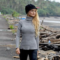 A woman with long blonde hair wearing a gray turtleneck sweater, black pants, and the Hand Crochet Martha Cap stands outdoors on a path lined with driftwood and trees in the background.