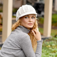 A woman wearing a gray ribbed turtleneck sweater and the Hand Crochet Martha Cap in white sits outdoors, gazing to her left. The blurred background features greenery and pale vertical columns.