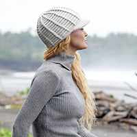 A person wearing a gray ribbed turtleneck sweater and the Hand Crochet Martha Cap—a handmade wool hat with a brim—stands outdoors, looking to the side. The background is blurred, showing greenery and a misty shoreline.