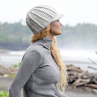 A person wearing a gray ribbed turtleneck sweater and the Hand Crochet Martha Cap—a handmade wool hat with a brim—stands outdoors, looking to the side. The background is blurred, showing greenery and a misty shoreline.