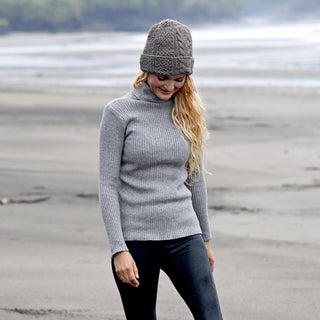 A person wearing the Hand Knit Inter hat, a gray ribbed turtleneck sweater, and dark pants stands on a sandy beach, looking down and smiling. The background is misty with distant greenery visible.