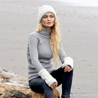 A person with long blonde hair, wearing a gray turtleneck sweater, white fingerless gloves, and the Hand Knit Inter hat, sits on a driftwood log at a sandy beach, gazing into the distance.