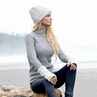 Wearing a cozy gray Hand Knit Inter hat, matching knit sweater, and gloves, a person sits on a log at the beach. The foggy background reveals sand and distant trees.