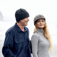 A man wearing the Hand Knit Nord Hat and a blue shirt walks beside a woman in a gray sweater and cap. They are outdoors on a foggy day near water or a beach, with the woman looking to her right and the man smiling.