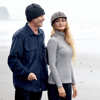 A man and a woman stand on a foggy beach. He wears a dark shirt and the Hand Knit Nord Hat, while she has long blonde hair, a gray sweater with hand knit wool details, and a newsboy cap. Both appear relaxed and casually dressed.