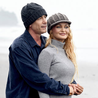 A man and woman stand on a breezy beach, both wrapped in cozy sweaters and wearing the Hand Knit Nord Hat, its fleece lining peeking out for extra warmth. The man stands behind the woman with his arms around her waist, as they gaze contentedly into the distance.
