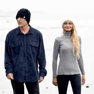 A man in a dark shirt and black cuffed beanie stands beside a woman dressed in a gray turtleneck, black pants, and the Hand Knit Nord Hat on a foggy beach. Both are looking slightly away from each other.