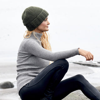 A person wearing a gray sweater, dark leggings, and the Hand Knit Timber Hat sits on a sandy beach, facing to the left. The background is mostly white and gray, with some sand and rocks visible.