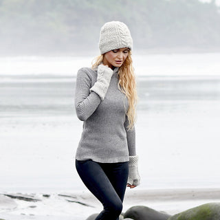 A person wearing a gray knit sweater, fingerless gloves, and a matching Hand Knit Inter hat walks along a foggy beach with water and trees in the background.