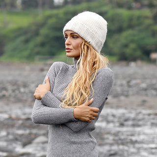 A person with long blonde hair wearing the Hand Knit Timber Hat and a gray ribbed sweater stands outdoors with arms crossed, looking to the side. The background is blurred, revealing rocky terrain and greenery.