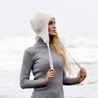A woman stands on a beach with ocean waves in the background, wearing a gray ribbed turtleneck sweater and the Hand Knit Snug Hat in cream. She holds the hat’s strings while looking off to the side.
