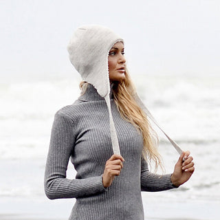 A woman stands on a beach with ocean waves in the background, wearing a gray ribbed turtleneck sweater and the Hand Knit Snug Hat in cream. She holds the hat’s strings while looking off to the side.