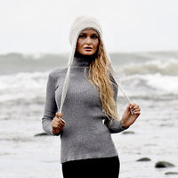 A person wearing a gray ribbed turtleneck sweater and the Hand Knit Snug Hat, a white hand-knit earflap hat with long ties, stands on a rocky beach with waves in the background. They have long, wavy blonde hair and are facing the camera.