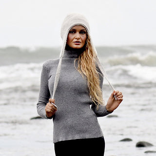 A person wearing a gray ribbed turtleneck sweater and the Hand Knit Snug Hat, a white hand-knit earflap hat with long ties, stands on a rocky beach with waves in the background. They have long, wavy blonde hair and are facing the camera.