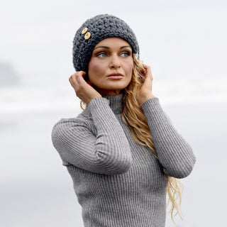 A woman with long blonde hair is standing outdoors in a misty environment, looking to the side, while wearing the Hand Crochet Acorn Hat and a gray ribbed turtleneck sweater.