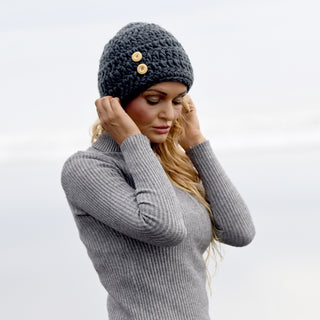 Wearing the Hand Crochet Acorn Hat, a person stands outdoors in an overcast setting, gently touching the sides of the cozy wool beanie adorned with two wooden buttons. They are also dressed in a ribbed gray sweater, looking down as the softly blurred background highlights the hat’s handcrafted detail.