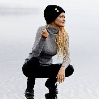 Wearing a hand crochet Acorn Hat, a person with long blonde hair, dressed in a gray turtleneck sweater, black pants, and black boots, squats on a reflective watery surface, looking to the right.