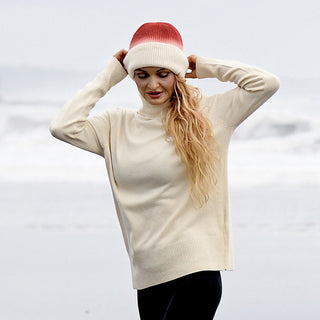 Wearing the Hand Knit Flow Hat in a red-and-white ombre style and a cream-colored turtleneck sweater, a person stands on the beach with waves in the background, looking down as they adjust their hat.