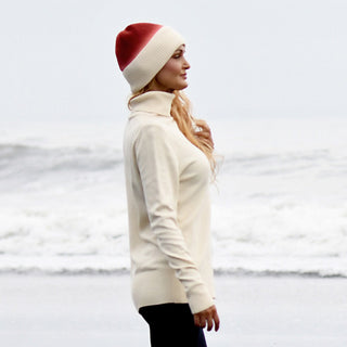 A person wearing the Hand Knit Flow Hat—a red and white 100% wool beanie—and a cream turtleneck sweater stands sideways on a beach, facing the ocean. Waves roll in under an overcast sky in the background.