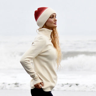 Wearing the Hand Knit Flow Hat, a woman in a cream turtleneck sweater stands in profile by the ocean, framed by waves and an overcast sky.