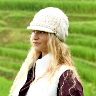 A person with long blonde hair wears the Broadway Hat and a white vest over a dark shirt, standing outdoors in front of green, terraced fields.