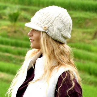 A person with long blonde hair wears the cream-colored Broadway Hat and a white vest over a dark top, standing outdoors before green, terraced fields.