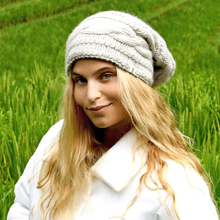 A person with long blonde hair in a Triple Braid Cable Slouch hand-knit beanie and a white coat stands outdoors before green grassy fields.