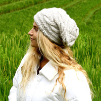 A person with long, wavy blonde hair is standing outdoors in a green field, facing to the left, wearing a white coat and the Hand Knit Triple Braid Cable Slouch beanie made from chunky merino wool.