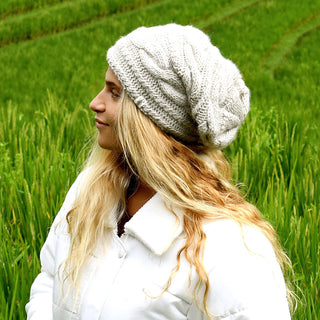 A person with long, wavy blonde hair is wearing a cream Triple Braid Cable Slouch beanie and a white puffer jacket while standing in a green field, facing to the left.