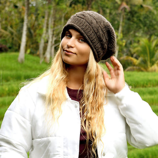 Wearing the Gateway Hat, a person with long blonde hair and a white jacket stands outdoors amid lush greenery, raising one hand near their lightweight wool hat lined with faux fur.