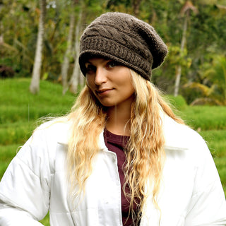 A person with long blond hair wears the Gateway Hat, a brown lightweight wool accessory handmade in Nepal, along with a white jacket, standing outdoors with blurred green trees and foliage in the background.