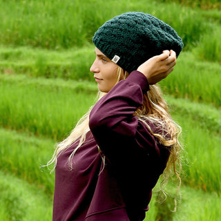 A person with long blonde hair wears a dark green Hand Knit Puff Beanie and a maroon sweater, standing in profile against a background of terraced green fields.