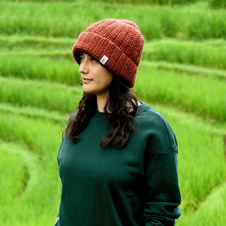 A person wearing a dark green sweatshirt and the Hand Crochet Chunky Hat stands outdoors in front of green, terraced fields, looking to the left.