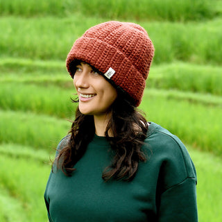 A person with long dark hair, wearing the Hand Crochet Chunky Hat in a reddish-brown hue and a green sweatshirt, stands outdoors in front of lush green grass or rice terraces, smiling and looking to the side.