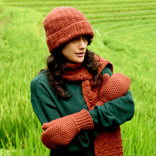 A person stands in a green field wearing the Hand Crochet Chunky Hat in a rust color, along with a matching crocheted scarf and mittens, and a dark green sweater. The individual has long, dark hair and is facing slightly to the left.