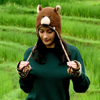 A person stands outdoors in front of green grass or fields, looking down and holding the braided strings of the Hand Crochet Grizzly Hat—a brown bear hat with adorable ears and face, hand crocheted from wool with a cozy sherpa lining—paired with a green sweater.