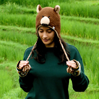 A person stands outdoors in front of green grass or fields, looking down and holding the braided strings of the Hand Crochet Grizzly Hat—a brown bear hat with adorable ears and face, hand crocheted from wool with a cozy sherpa lining—paired with a green sweater.