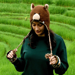 Wearing the Hand Crochet Grizzly Hat—a hand-crocheted wool hat with cozy sherpa lining—a woman stands outdoors in a green field, looking down and holding the braided strings of her bear-themed hat. She pairs it with a green sweater and colorful patterned fingerless gloves.